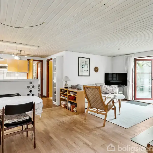 Stue med lyse trægulve indeholder spisebord og stole til venstre, sofahjørne med lænestol og fladskærms-TV til højre. Store glasdøre åbner ud til en terrasse.