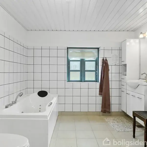 Et hvidt badeværelse med et badekar står ved siden af en toilet. En bruser ses ved et vindue med et brunt håndklæde hængende. Der er også et spejl og en vask.