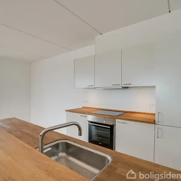 Et moderne køkken med en indbygget vask i en træbordplade, der strækker sig over en hvid skabsvæg med integreret ovn. Rummet er lyst med et vindue i baggrunden.