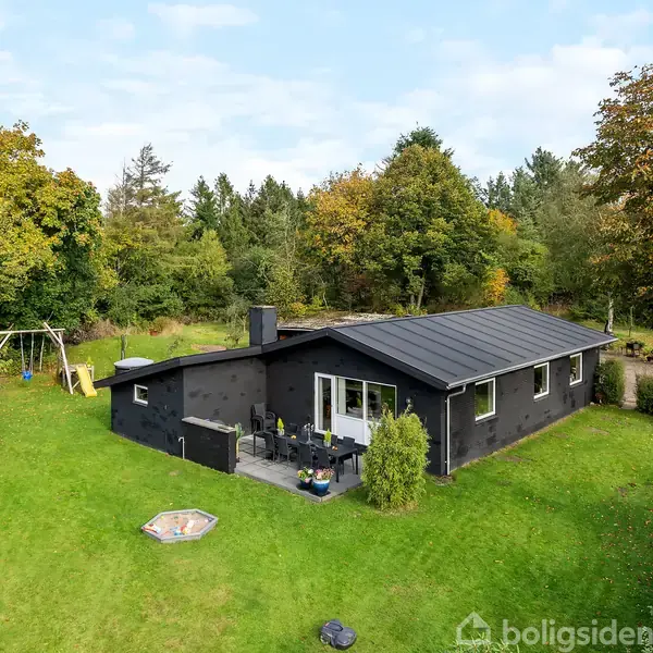 En sortmalet bungalow står på en grøn græsplæne med terrasse og have. En sandkasse er i forhaven. Træer omgiver huset, og en gynge står til venstre.