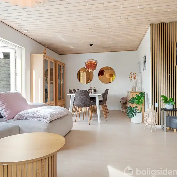 Lys stue med beige sofa og rundt sofabord, spisebord med stole i baggrunden omgivet af planter og dekorative elementer. Store vinduer slipper lys ind.