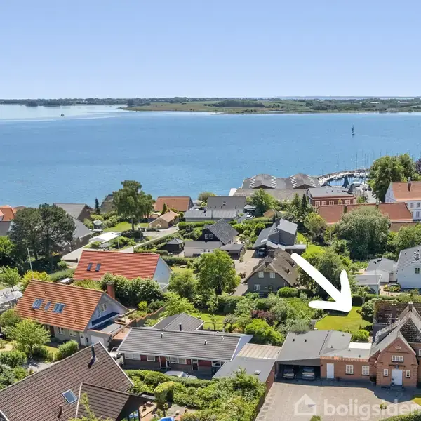 En pil peger mod et hus med ovenlysvinduer, omgivet af andre boliger og græs, tæt ved en havn. I baggrunden ses havet og en skovbeklædt kystlinje.