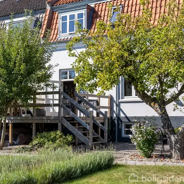 Hus med rød tagstenstag og tilhørende træterrasse, hvor en trappe fører ned til haven. To store træer står i den grønne have, omgivet af buske og græs.