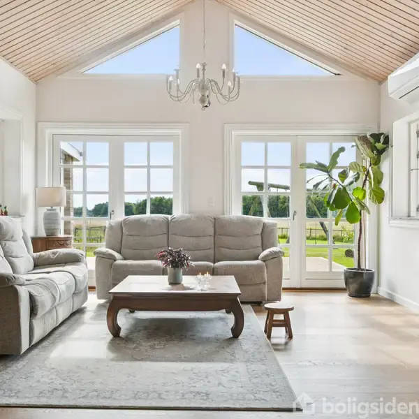 Sofaer står stille i en lys stue med højt til loftet. Store vinduer åbner op til grøn natur. En lysekrone hænger over et sofabord, og en plante står ved døren.