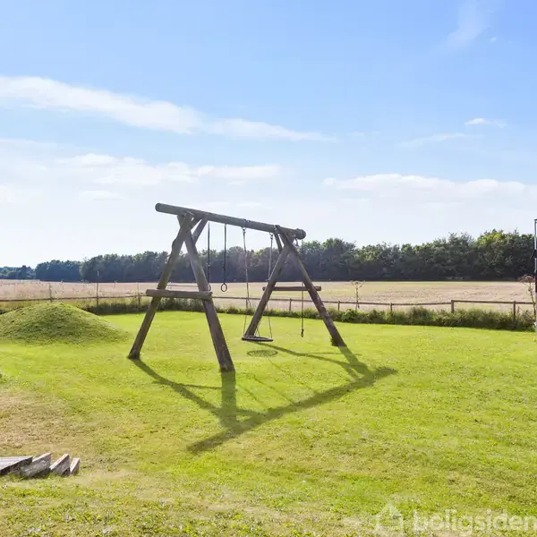 Gyngestativ med to gynger står på en grøn græsplæne i en indhegnet have. Omkring er der åbent landskab og en skovkant i baggrunden. Solrigt vejr.