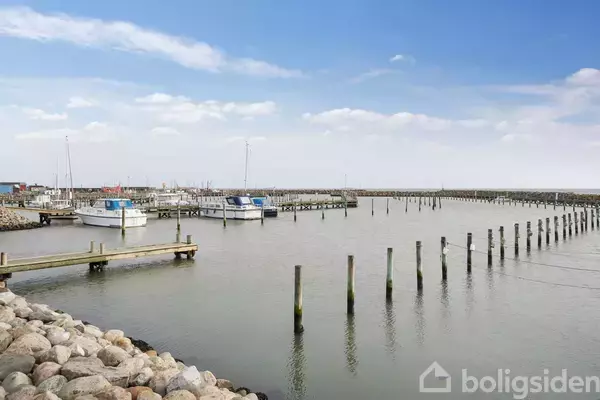 Flere både ligger til kaj i en stille havn omgivet af moler og en rolig, blå himmel. Rundt omkring er der stenmoler, der danner beskyttelse mod vandet.