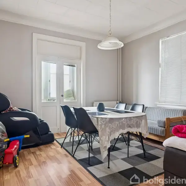 Stue med spisebord dækket med dug og omgivet af fem stole. Til venstre står en lænestol, og i hjørnet nær vinduet er en sofa. Legetøj og tæppe på gulvet.