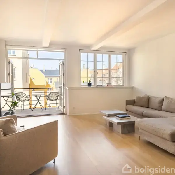 Stue med beige sofa og lænestol, vender mod en åben altandør. Udenfor ses en lille altan med bord og stole. Lyse vægge og gulv dominerer rummet.