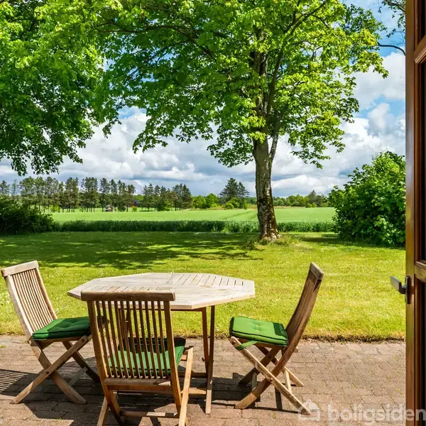 Et rundt træbord med fire stole står på en terrasse. Sollys skinner på græsplænen omgivet af frodig natur og træer.