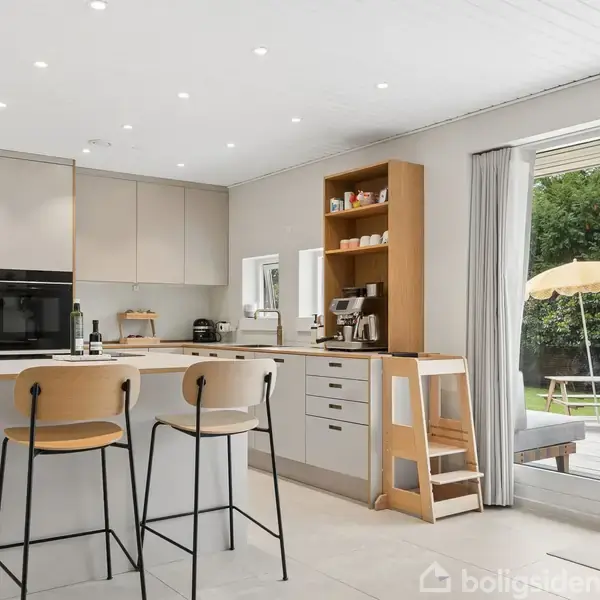 Et moderne køkken med to høje stole omkring en ø. Lys indretning med indbyggede skabe, åben hylde, og en stor glasdør åbner til en terrasse med grøn udsigt udenfor.
