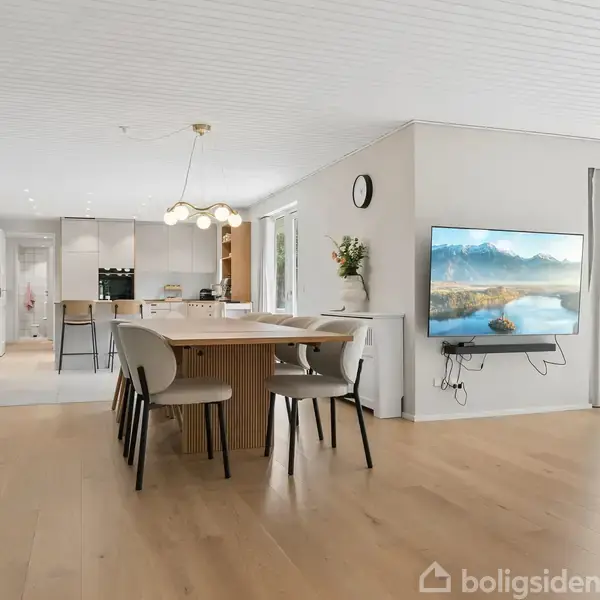 Spisestue med rundt bord og seks stole i moderne, lyst interiør. Vægmonteret fladskærm viser landskab. Store vinduer lysner rummet. I baggrunden ses et køkkenområde.
