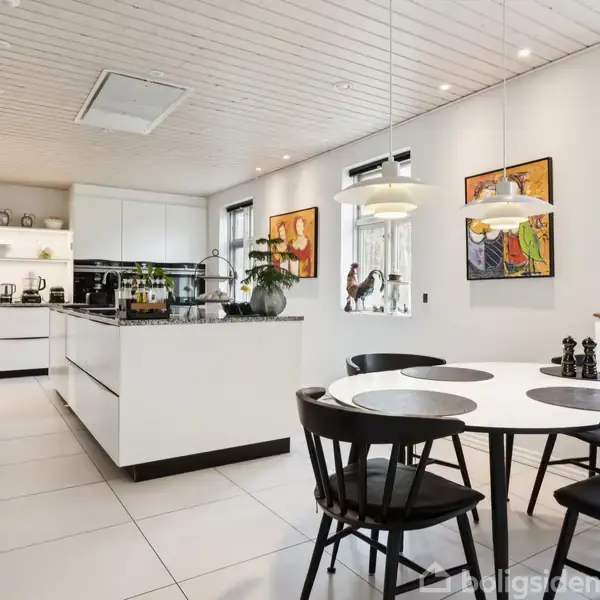 Et moderne køkken med en central ø, hvorpå der står planter. En rund spisebord med sorte stole står i rummet. Lyse fliser og skandinavisk design præger indretningen.