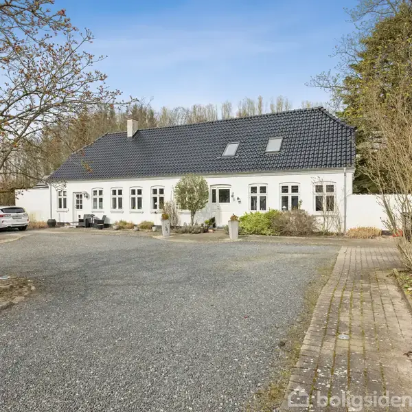 Hvidt hus med sort tag står stille i en gårdsplads. Omkring er der parkerede biler, træer uden blade og en brolagt sti.