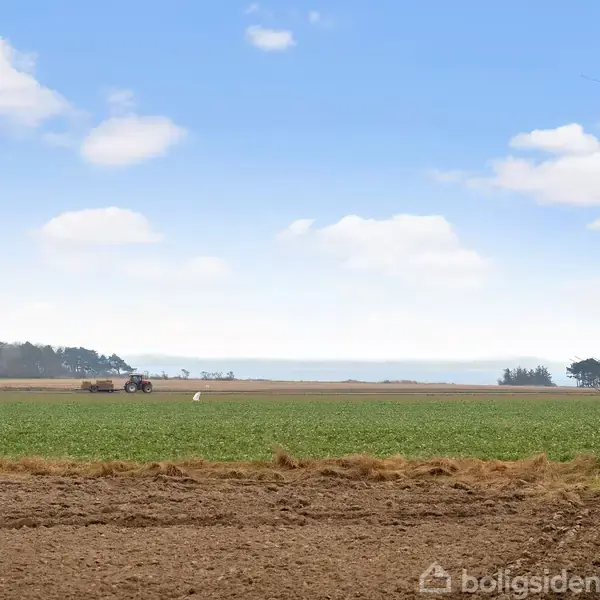 Traktor bevæger sig langsomt over en stor mark, omgivet af åbne, grønne felter og skov. I baggrunden ses havet under en blå himmel med spredte skyer.