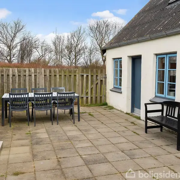 Et bord med seks stole står på en flisebelagt terrasse ved siden af et hvidt hus med blå vinduer og dør. Bagved ses et hegn og træer.