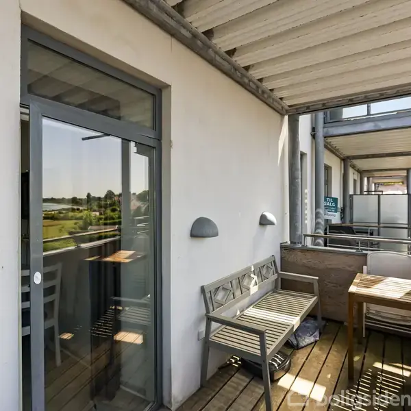Balkon med træmøbler, inklusive en bænk og et bord, står ved siden af en glasdør. Balkon rækværk og udsigt over grønt landskab skaber en lys og åben atmosfære.