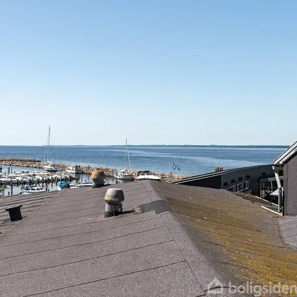 Tagtag med udsigt over en marina, hvor både ligger ved molen. I baggrunden ses havet under en klar blå himmel.