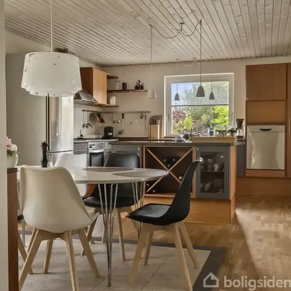 Rundt spisebord med sorte og hvide stole i et moderne køkkenmiljø. Køkkenet har en metalemhætte, træfarvede skabe og hylder samt en bordplade ved et vindue med udsigt til grønne træer.