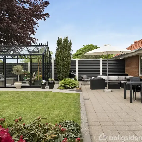 Drivhus i haven står ved siden af en terrasse med havemøbler og parasol. Rødblomstrende planter grænser op til en velplejet græsplæne. Murstenshus ses i højre side.