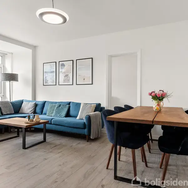En blå sofa står med puder i en stue. Et træbord med blomster står foran, omgivet af stole. Rummet er lyst med vindue og billeder på væggen.