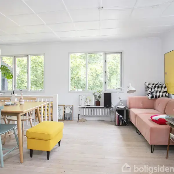 En stue med en lyserød sofa ved siden af et lille bord. En spisebord med pastelstole og et gult skammel står til venstre. Store vinduer giver udsigt til grønne træer.