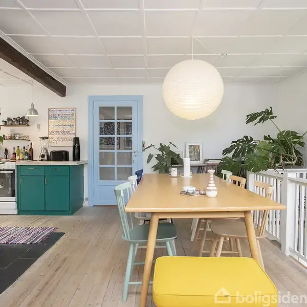 Rummeligt spisestueområde med et langt træbord og farverige stole. En stor rund lampe hænger over bordet. Til venstre ses en brændeovn og et åbent køkken med grønne skabe. Grønne planter dekorerer rummet.
