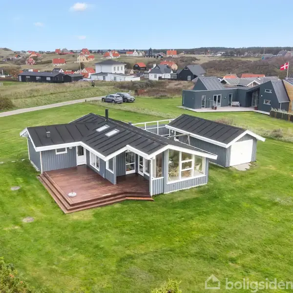 Et gråblåt sommerhus med flere vinduer står på en velplejet græsplæne. En veranda er vedhægt på huset. I baggrunden ses andre huse og en bil på en landevej.