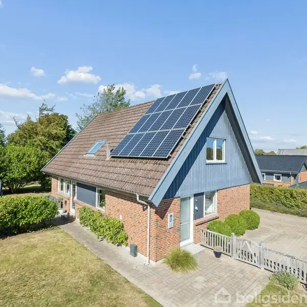 Et hus med solpaneler på taget står i solen. Omkring det er en frodig have og en flisebelagt indkørsel. Flere buske omkranser ejendommen.