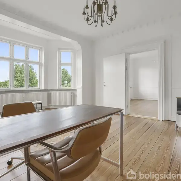 Et lyst skrivebord med stole står placeret på et trægulv i et rum med store vinduer og en lysekrone. Rummet er forbundet til et tilstødende værelse gennem en åben døråbning.
