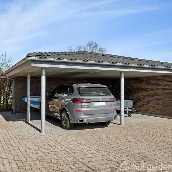 En sølvfarvet bil holder parkeret under en carport ved siden af et blåt kajak. Carporten er lavet af mursten og træ, omgivet af en flisebelagt gårdsplads.