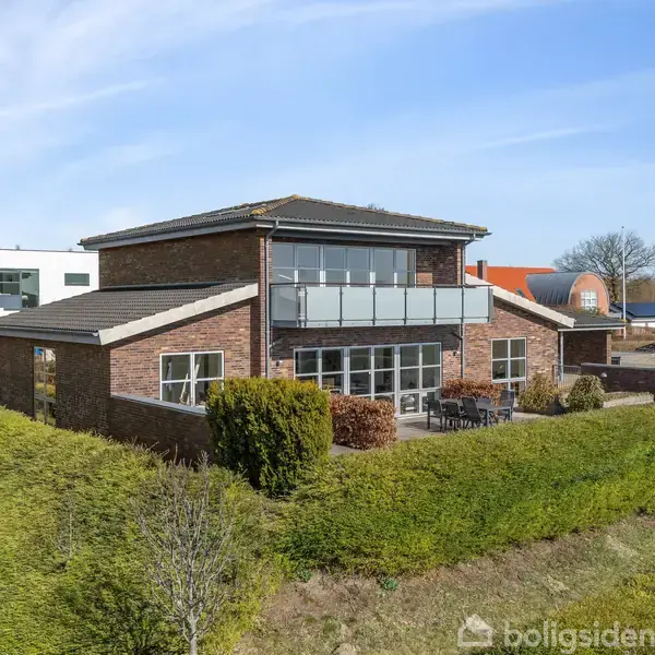 Et moderne murstenshus har store vinduer og en balkon med glasværn. Det står i et boligområde omgivet af hække og andre huse.