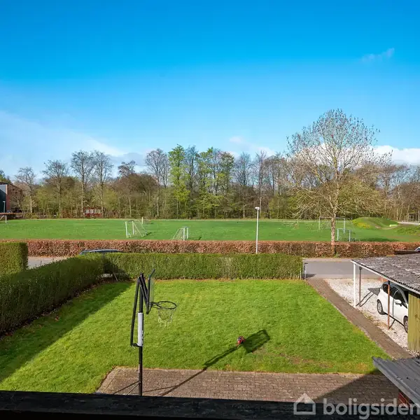 En grøn have med en tørresnor strækker sig mod en større græsplæne med fodboldmål. Træer omkranser området under en klar blå himmel.