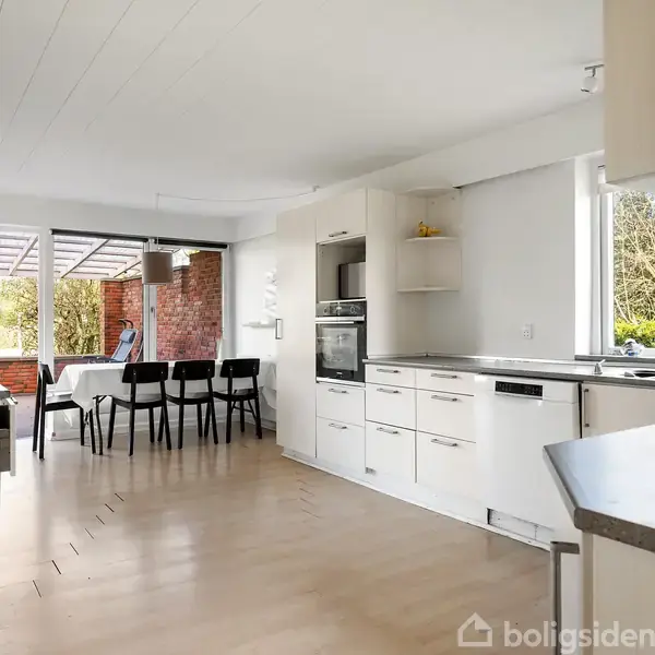 Hvidt køkken i moderne stil, udstyret med ovne, skabe og en vask. Spisebord med sorte stole ved vinduet. Udsigt til en terrasse og grønne træer udenfor.