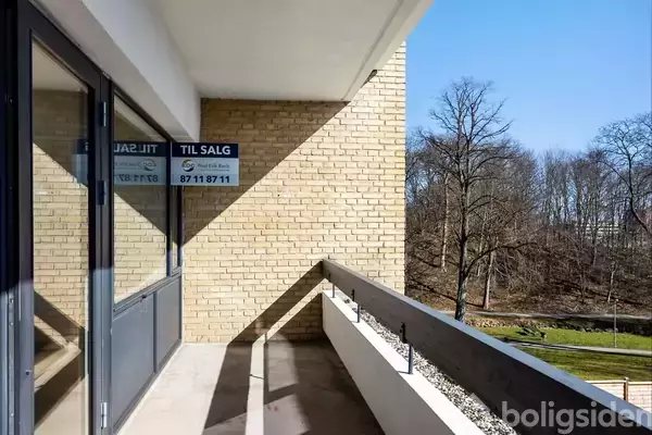 En altan med murstensvæg og glasdøre viser et "TIL SALG" skilt med telefonnummer 87 11 87 11. Baggrunden har træer og en klar blå himmel.