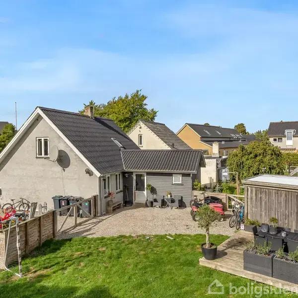 Et gråt hus med sort tag står i en have med grøn græsplæne. Flere cykler står udenfor ved en grusvej, omgivet af andre huse i nabolaget.