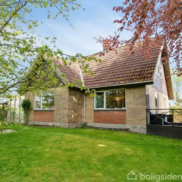 Et murstenshus med skråt tag står i en frodig have med grøn græsplæne og træer rundt omkring. Huset har store vinduer og en lille terrasse ved siden af indgangen.