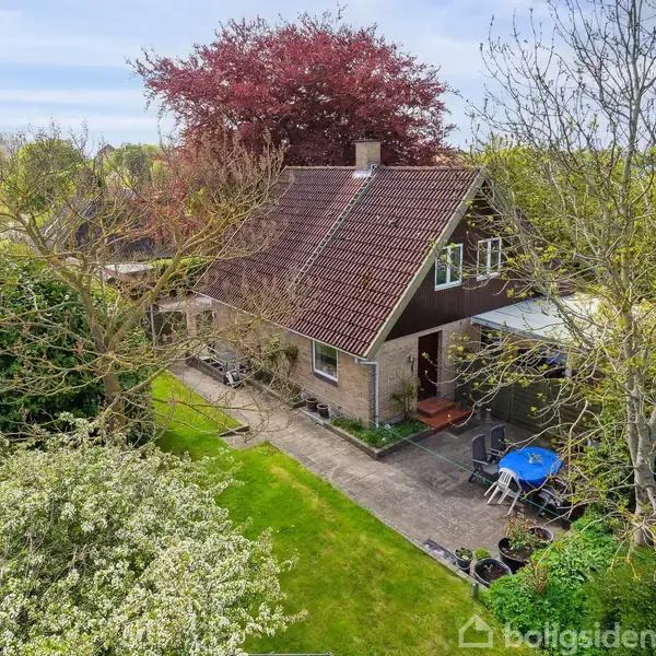 Et to-etagers hus med skråt tag står omgivet af en frodig have. En terrasse med blå bord og stole ses foran, mens træer og buske omgiver ejendommen.