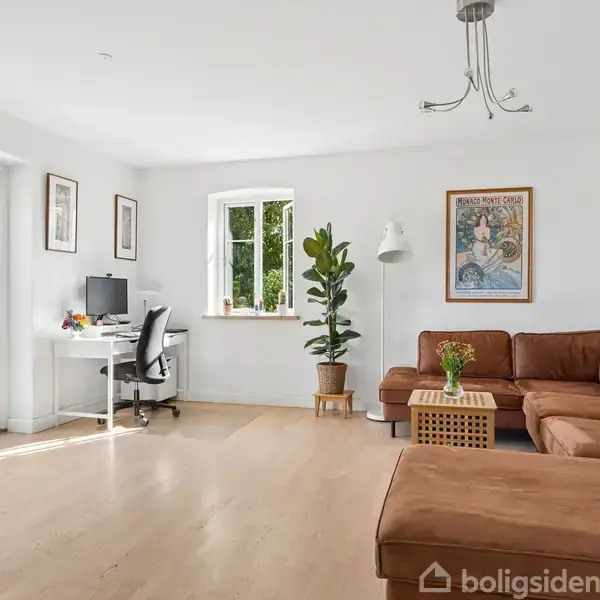 Brun hjørnesofa står i lys stue med billeder på væggene, ved siden af et skrivebord med computer. Store vinduer og døre åbner mod en grøn have.