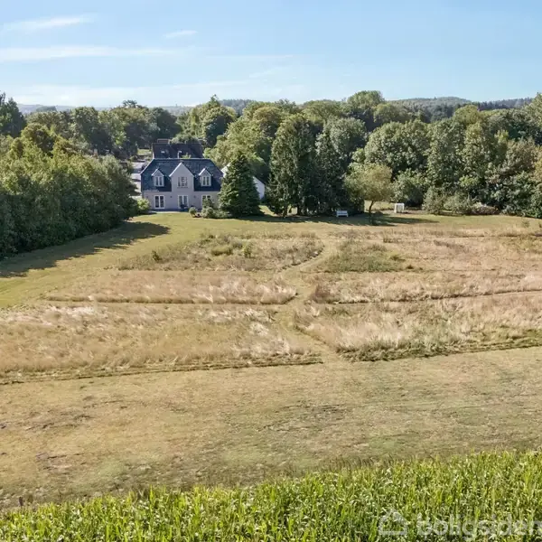 Et hvidt hus med mørkt tag står i baggrunden omgivet af buske og træer. Foran huset ligger en stor, åben mark med græs i mønstre, omgivet af grønt landskab.
