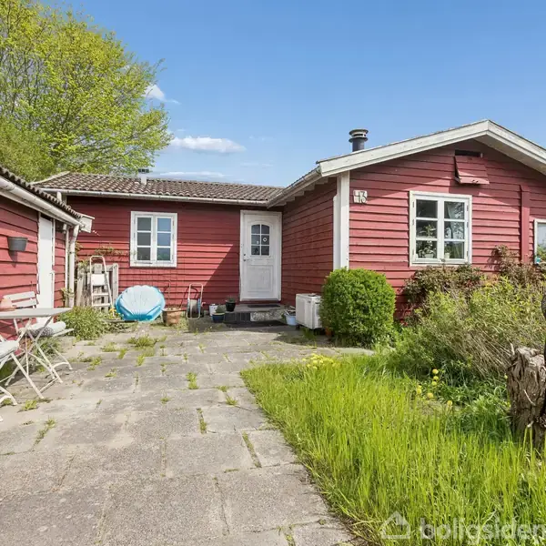 Rødt træhus med hvide vinduer og dør stående stille. En gårdsplads med fliser og græs til højre. To hvide metalstole og bord placeret langs venstre mur. Træer i baggrunden.
