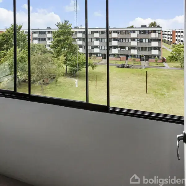 Balkon med glasvægge udenfor en bolig, ingen aktivitet, udsigt over grønne områder med træer og en lang, lav beboelsesbygning i baggrunden. Klar himmel med nogle skyer.