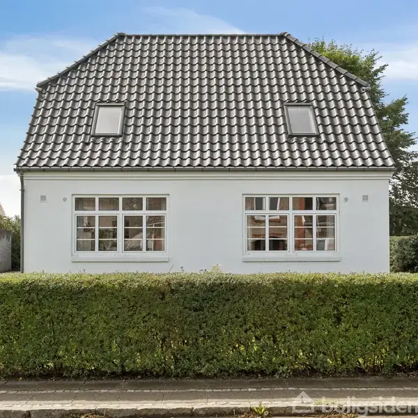 Et hvidt hus med mørkt tegltag står bag en lav hæk. Sollys oplyser facaden, hvor to vinduer og to tagvinduer er synlige. Fjern omgivelser er træbevoksede.