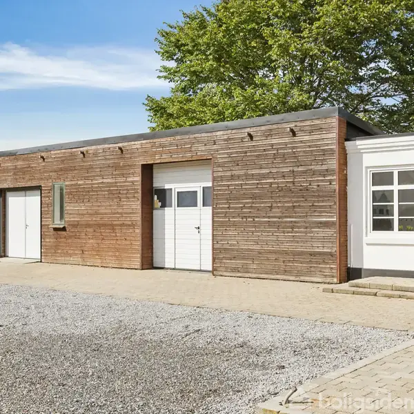 En træbeklædt garagebygning med to hvide porte, placeret ved siden af en hvid murstensbygning. Beliggende på en grusbelagt gårdsplads, omgivet af en grøn hækkant og træer.