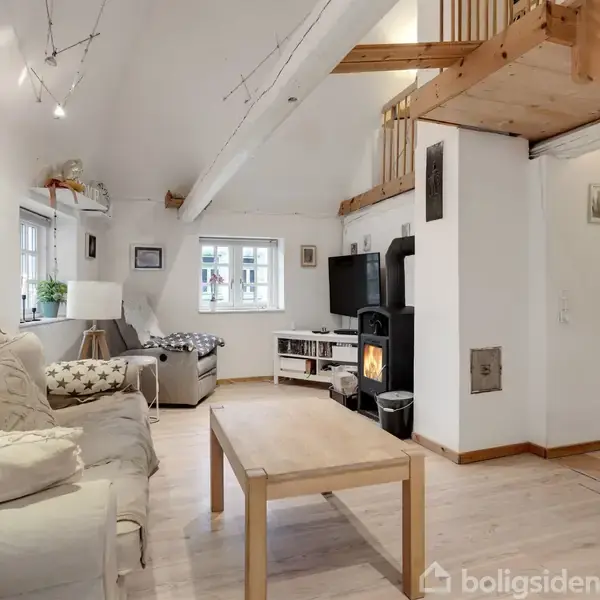En lys stue med sofa og sofabord i forgrunden. En brændeovn er tændt i hjørnet, og en trappe fører op til en hems. Vinduerne giver naturligt lys.