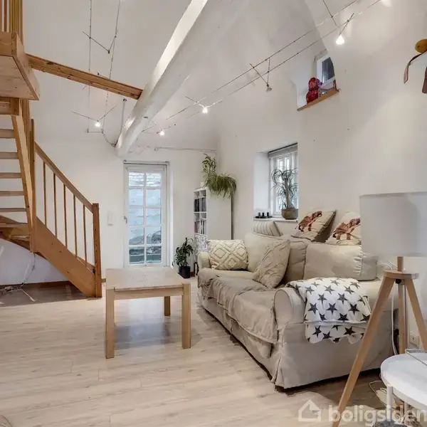 Sofa i stue står ved væg under skråt loft, dekoreret med lyskæder. Trætrappe i hjørne fører opad. Værelset har lyst trægulv, vinduer og en glasdør med planter omkring.