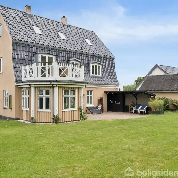 Et stort, beige hus med sort tag har en balkon og terrasse. Haven er rummelig med en grøn græsplæne, og der er et lille, tilstødende skur i baggrunden.