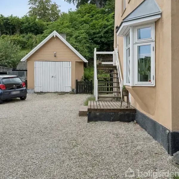 Et hus med en tilknyttet trappe og en grusbelagt forplads. En garage med hvid dør står ved siden af, og en bil er parkeret tæt på.