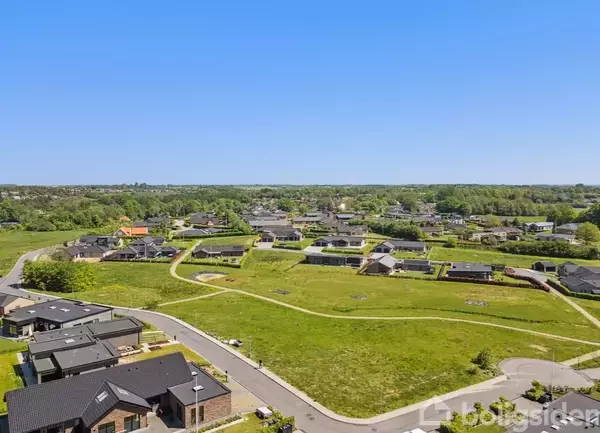 Boligområde set fra luften, hvor moderne huse står langs snoede veje omgivet af grønne marker og træer. Det er en klar dag med blå himmel.