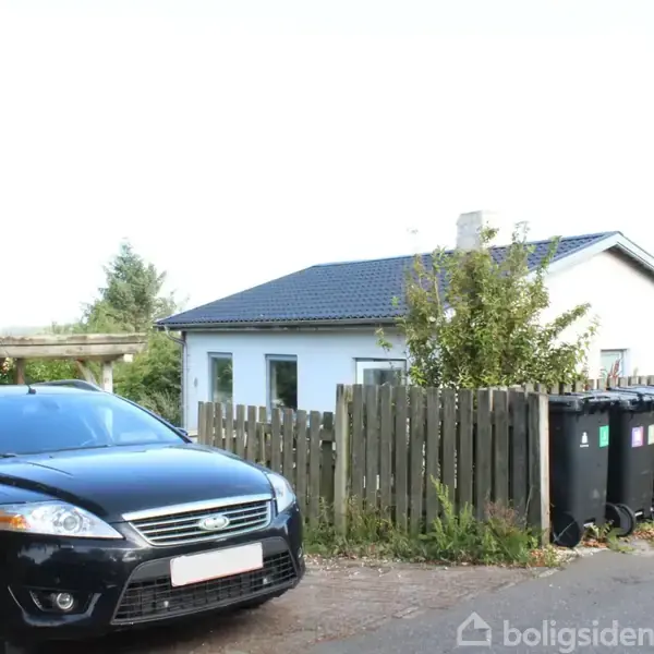 En sort bil holder parkeret foran en hvid bygning med blå tag. Ved siden af huset står en række affaldsbeholdere på en asfalteret vej, omkranset af et træhegn.