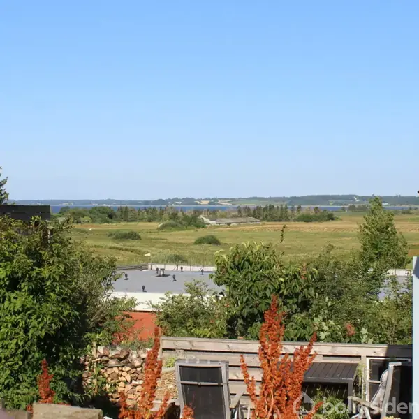 Hus i forgrunden med have fyldt med grønne planter og brune blomster, beliggende for enden af en træterrasse. I baggrunden ses en udsigt over åbne marker og en sø under en klar blå himmel.